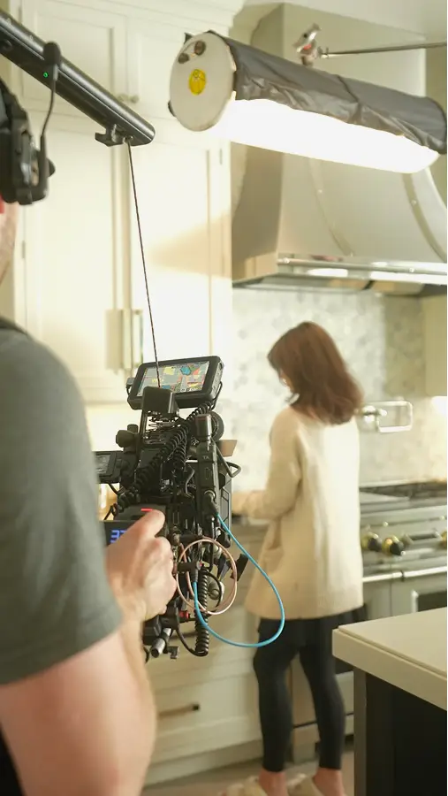 Ben filming a woman in a kitchen using a professional video camera with a large light source above.