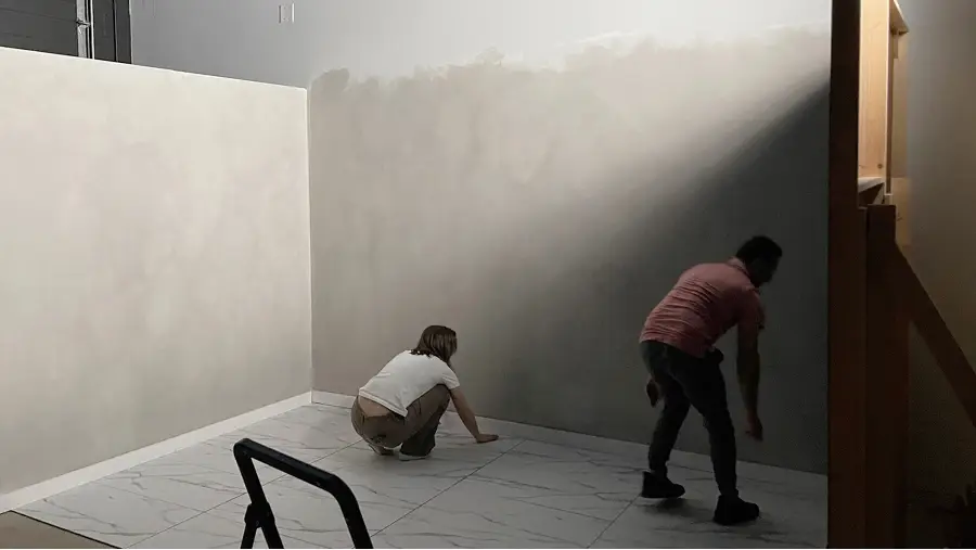 Emma and Achi crouching on a tiled floor inside The CAVE Studio with plain beige and gray walls under dim lighting.
