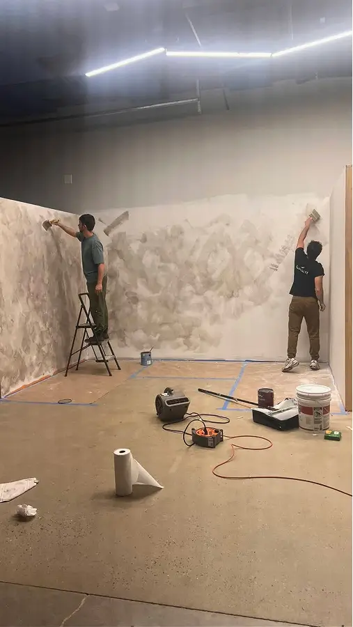 Team painting a large wall with a textured beige pattern in The CAVE Studio, one standing on a ladder and the other on the floor.