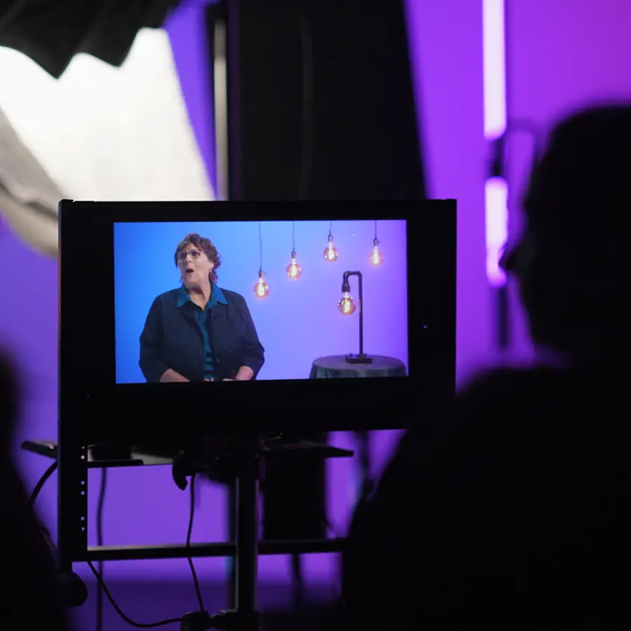 Monitor screen showing a woman speaking on a set in The CAVE Studio with hanging light bulbs, purple background and silhouette of a person watching.