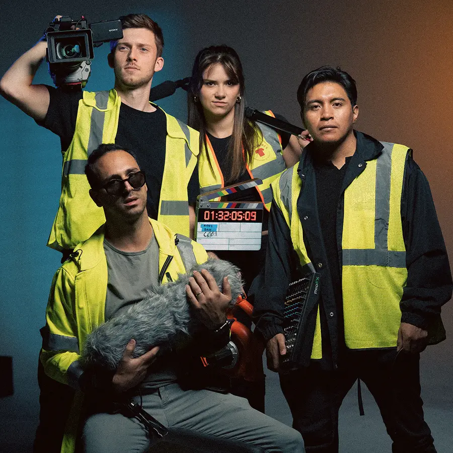 Crew wearing yellow safety vests, holding camera, clapperboard, microphone, and audio mixer.