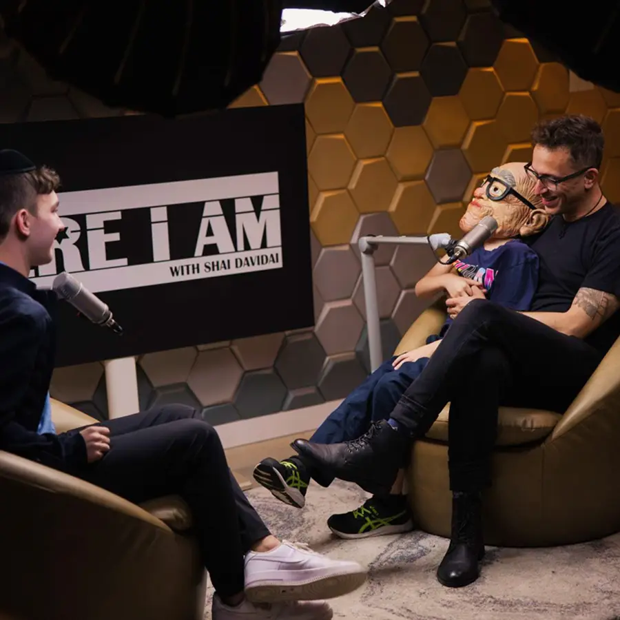 Two men in a podcast studio in The CAVE Studio, one holding a puppet with glasses and a microphone, smiling and interacting.