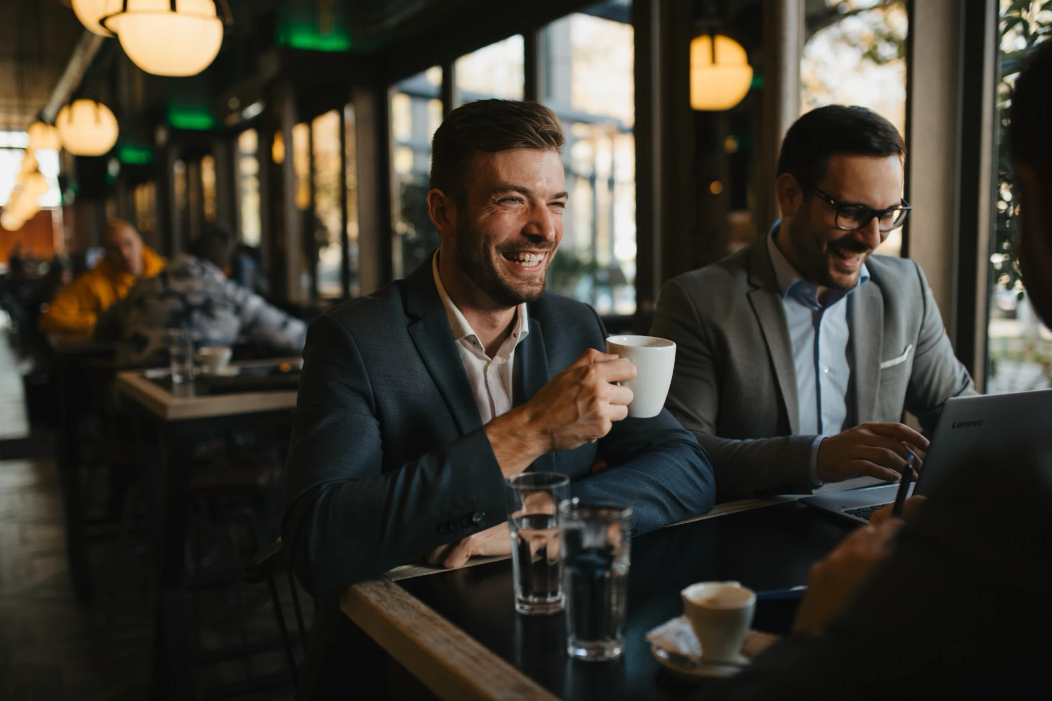 Two men in suits smiling and drinking coffee while working on a laptop in a cafe.