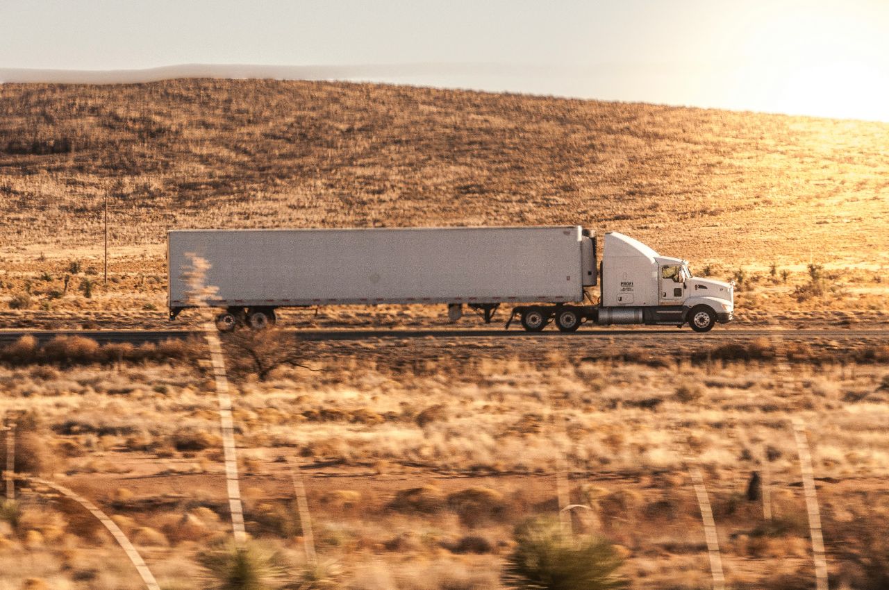 White semi-truck hauling a box trailer across a desert highway at sunset, transporting freight through open rural terrain.