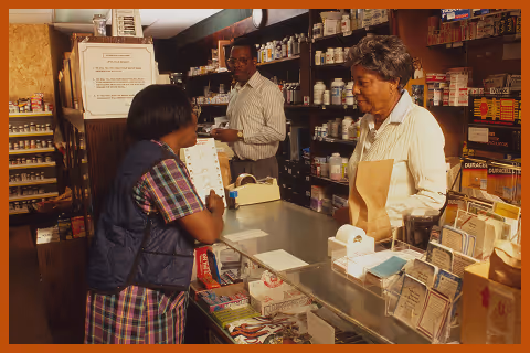 Customer at the counter of a pharmacy talking to a female pharmacist with a male assistant in the background.