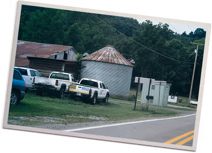 Parked vehicles alongside a rural road next to a rusted grain silo and wooden building with forested hills in the background.