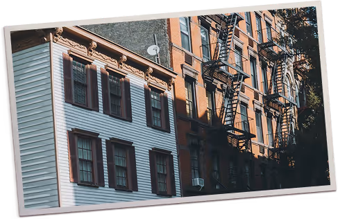 Side view of adjacent buildings with white siding and dark shutters next to a red brick building featuring black metal fire escapes.
