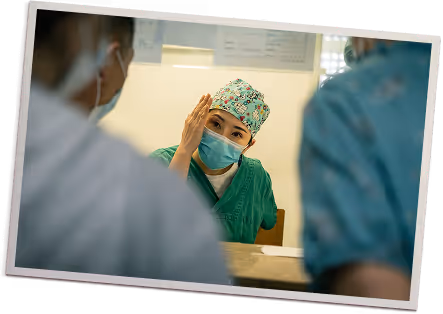 Medical professional in green scrubs and patterned cap adjusting face mask while talking to two people.