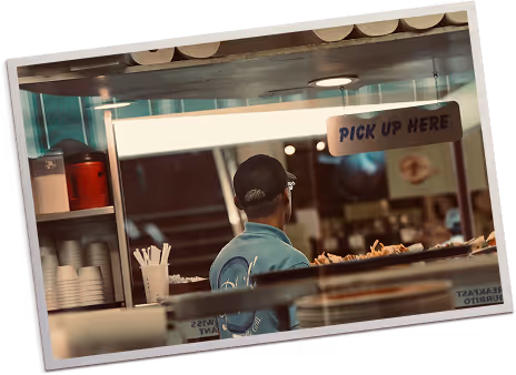 Person wearing a cap and blue shirt standing behind a counter at a food pickup window with a 'PICK UP HERE' sign.