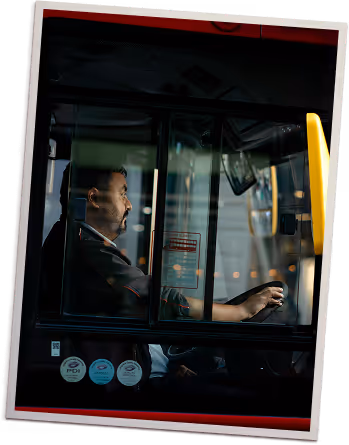 Bus driver seated at the wheel inside a bus cabin, visible through the glass window.