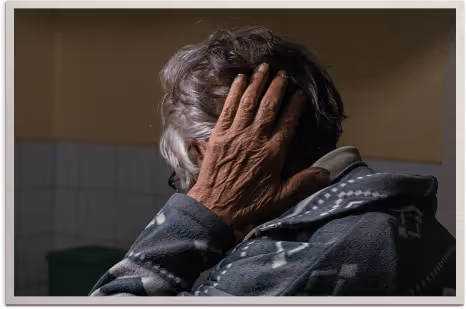 Elderly person with gray hair covering their face with a wrinkled hand, wearing a patterned jacket.