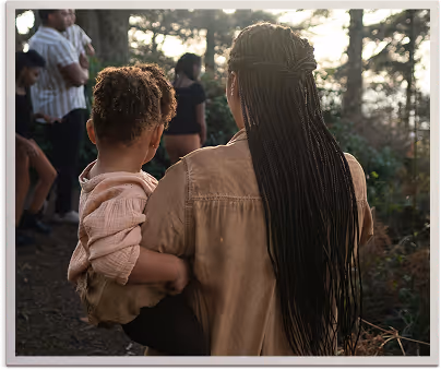 Woman with long braided hair carrying a toddler while walking outdoors among a group of people.
