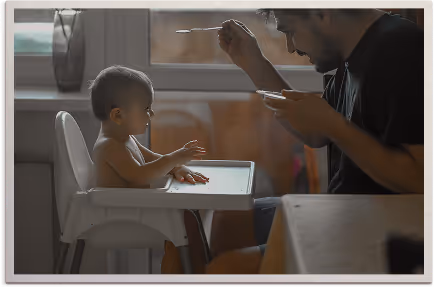 Father feeding a baby seated in a high chair by a window.