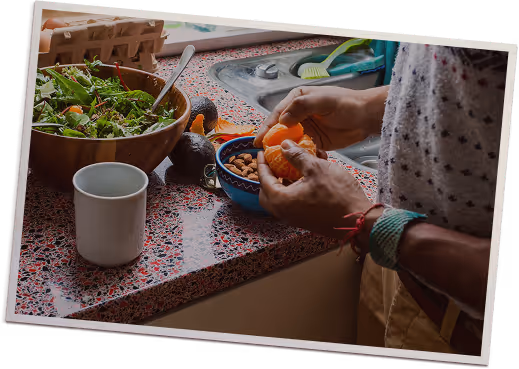 Person peeling an orange next to a bowl of salad, avocado, almonds, and a white mug on a kitchen counter.