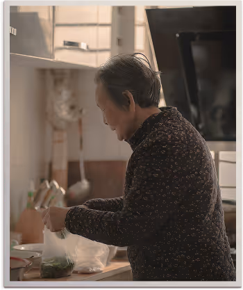 An elderly woman preparing food in a kitchen, placing greens into a plastic bag on the counter.