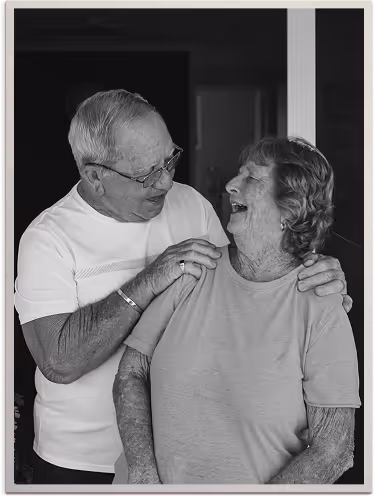 Elderly man wearing glasses affectionately holding and smiling at an elderly woman who is laughing.