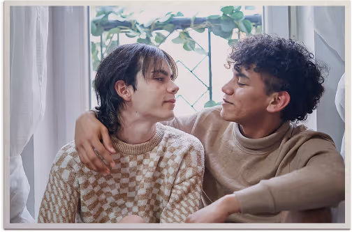 Two young people sitting close together by a window, one with an arm around the other, sharing a warm, affectionate gaze.