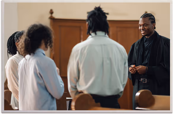 A man in a black graduation gown smiling and speaking to three other people in a wooden-paneled room.
