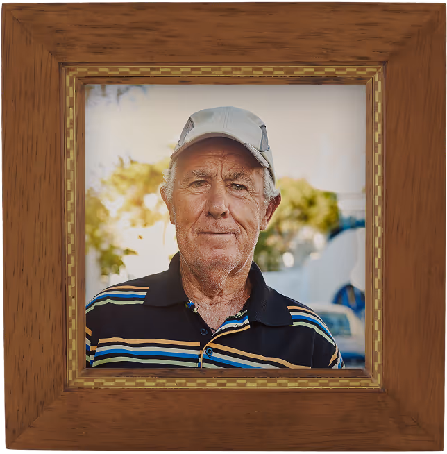 Portrait of an older man wearing a light gray cap and a striped polo shirt framed in a wooden picture frame.
