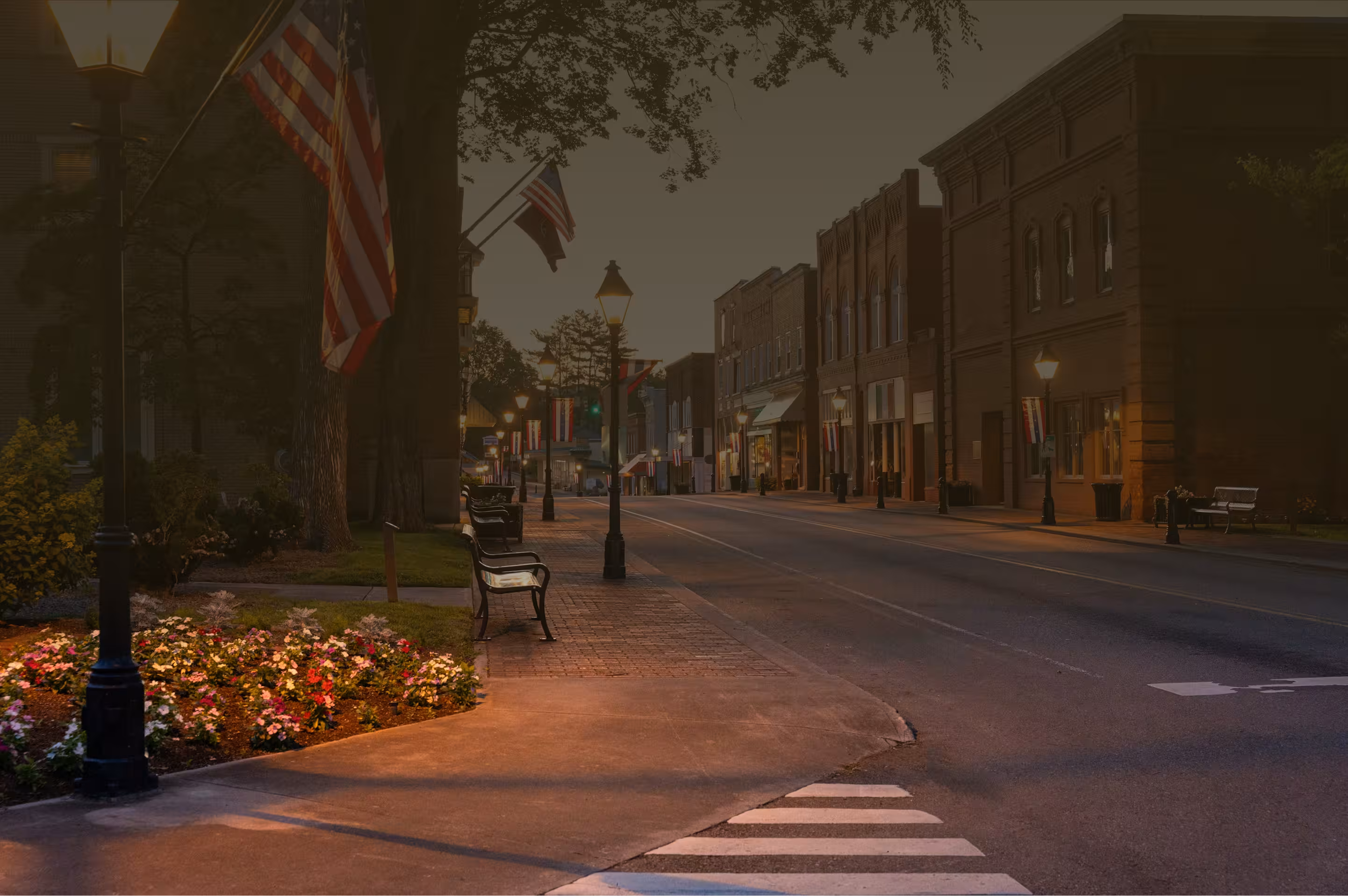 Quiet small-town street at dusk with American flags, benches, lit street lamps, and brick buildings.