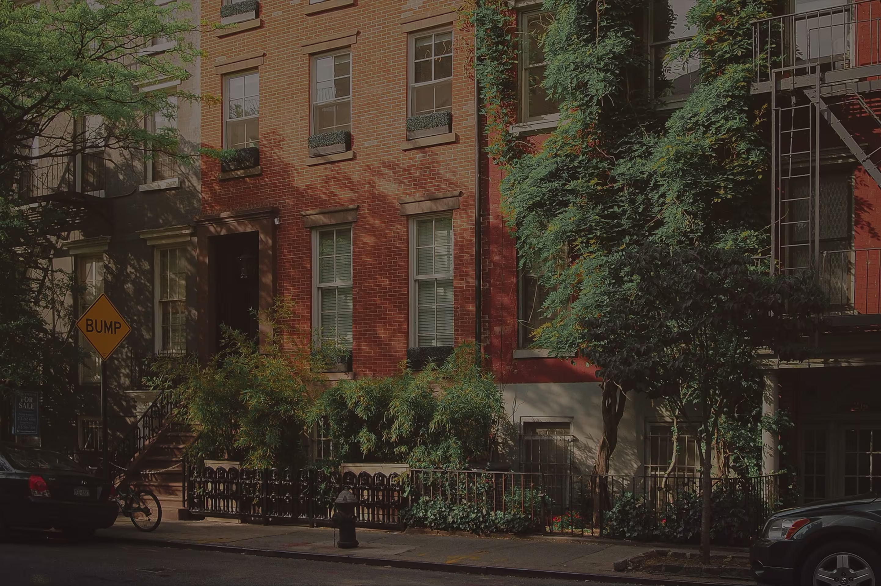 Tree-lined street with brick townhouse, green shrubs, parked cars, and a yellow BUMP road sign.