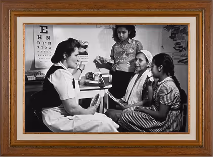 Black-and-white photo of a healthcare worker conducting an eye exam with an eye chart behind, speaking with three women seated and standing nearby.