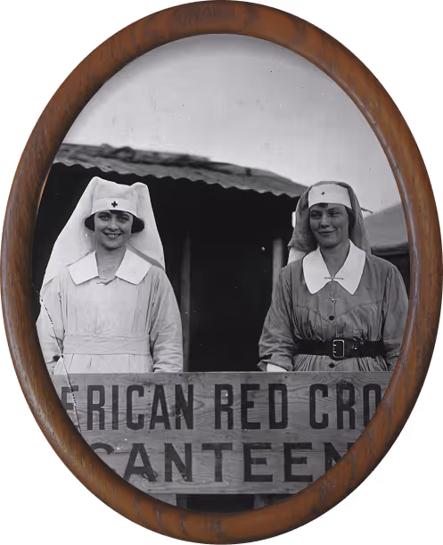 Two smiling nurses in Red Cross uniforms standing behind a sign that reads 'American Red Cross Canteen' framed in an oval wooden frame.