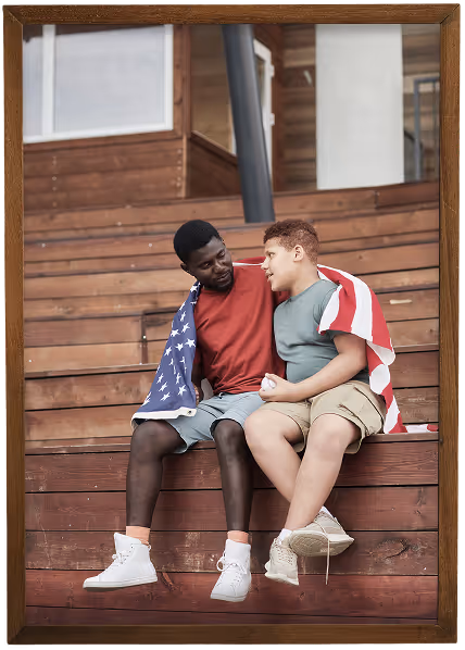 Two boys sitting on wooden steps, wrapped together in an American flag, looking at each other and smiling.