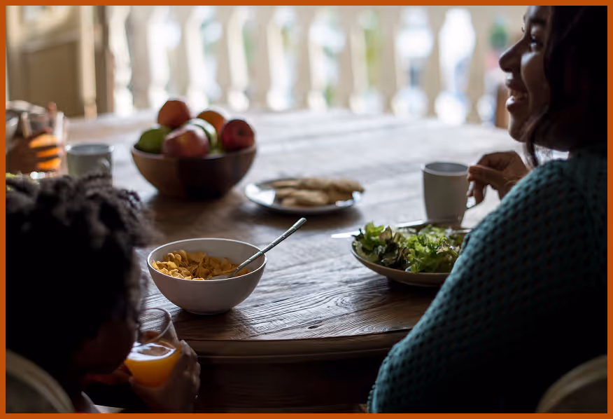 Two people sitting at a wooden table with bowls of cereal, salad, a plate of crackers, a cup, and a bowl of fruit.