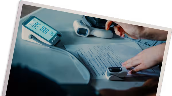 Person recording data on paper with a pen next to a digital blood pressure monitor, pulse oximeter, and thermometer on a table.