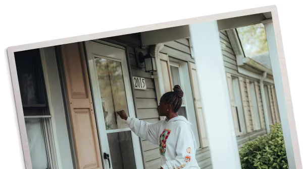 Woman wearing a white hoodie and mint green pants standing at a gray house door, knocking.
