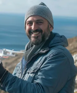 Man wearing a gray knit hat and blue jacket smiling outdoors with a drone in his hand near a body of water.