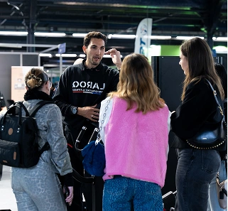 Four people standing indoors engaged in conversation, with one man wearing a black LAROO sweatshirt explaining something.