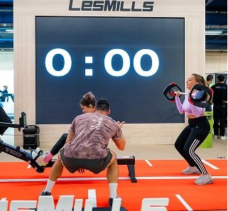 Two people exercising with weights in front of a large LES MILLS timer display showing 0:00.