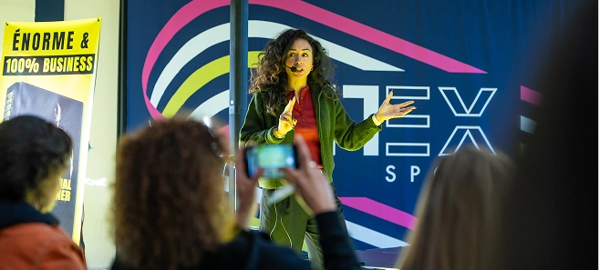 Woman speaking and gesturing during a presentation with audience members watching and one filming with a smartphone.