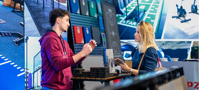 Man in a red hoodie explaining something to a blonde woman at a counter with various small black boxes.