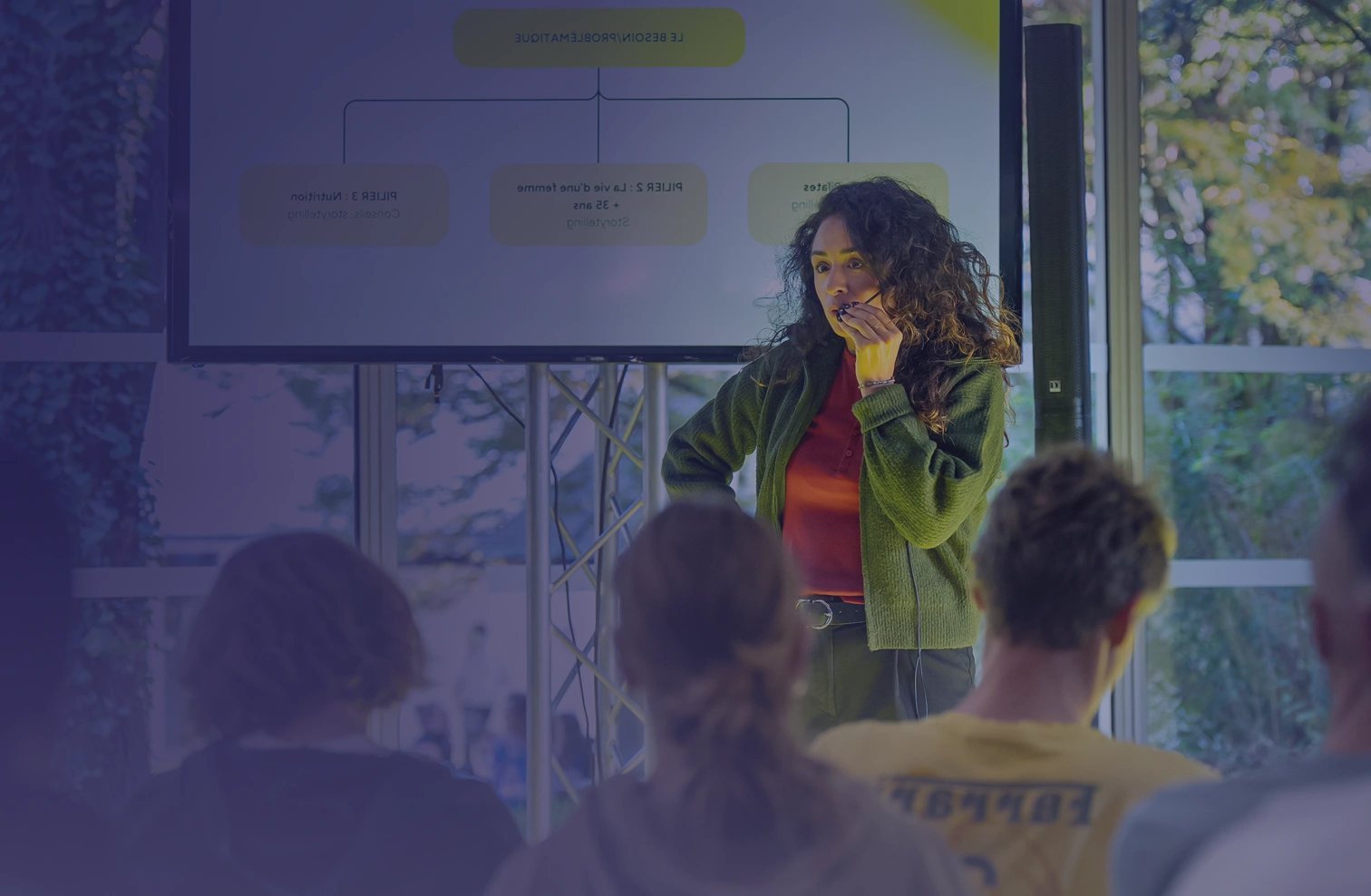 Woman with curly hair speaking into a microphone during a presentation in front of an audience.