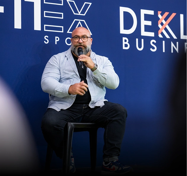 Bald man with glasses speaking into a microphone while sitting on a black stool against a blue backdrop with white text.