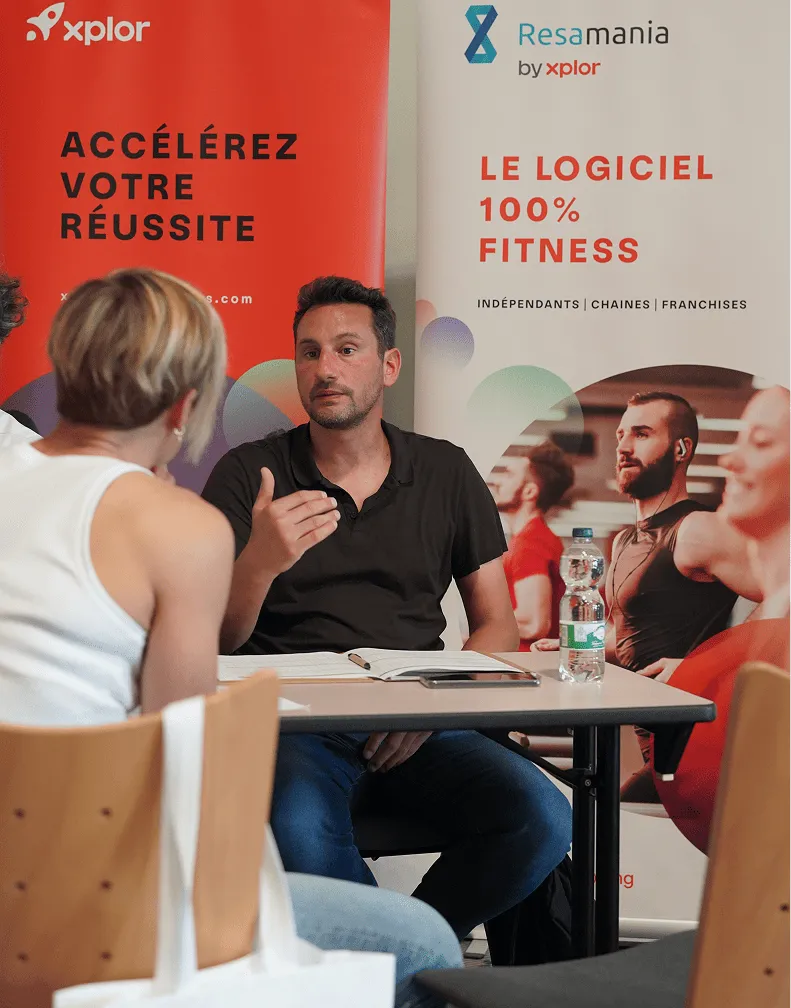 Man in black shirt explaining something to a woman with short blonde hair seated at a table with open notebooks and a water bottle, against banners promoting Xplor fitness software.