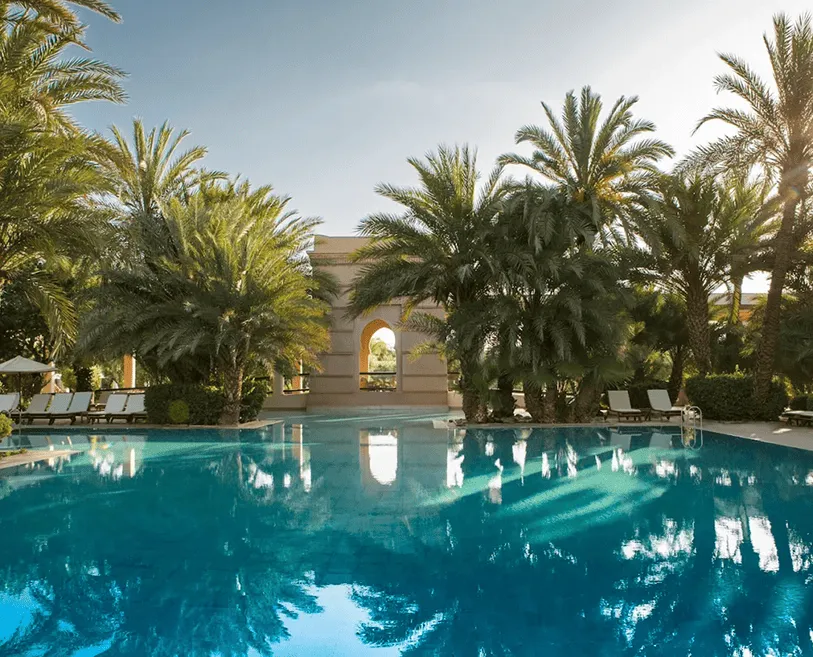 Outdoor swimming pool surrounded by palm trees and lounge chairs with sunlight reflecting on the water.