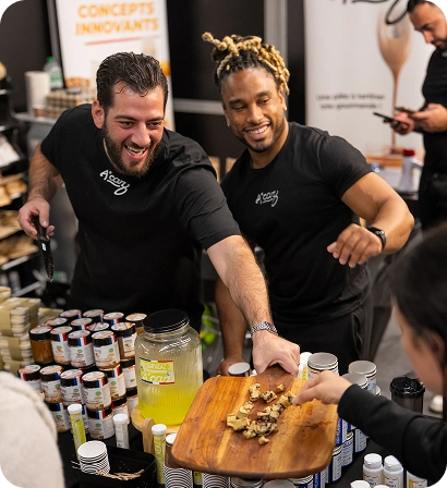 Two men in black shirts offering food samples on a wooden board at a stall with jars and bottles on display.