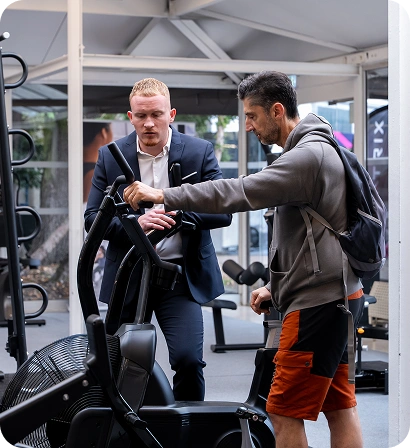 A gym trainer instructing a man in a hoodie and orange shorts on how to use an exercise bike.