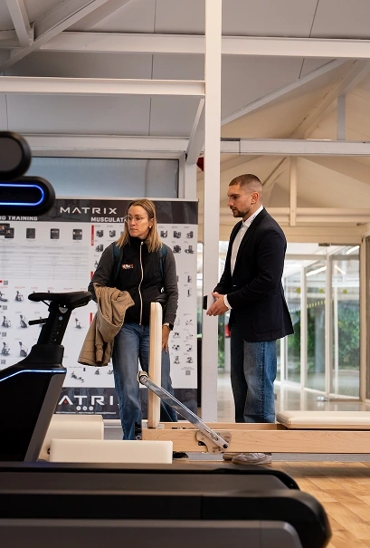 Two people standing in a gym near workout equipment, engaged in conversation.