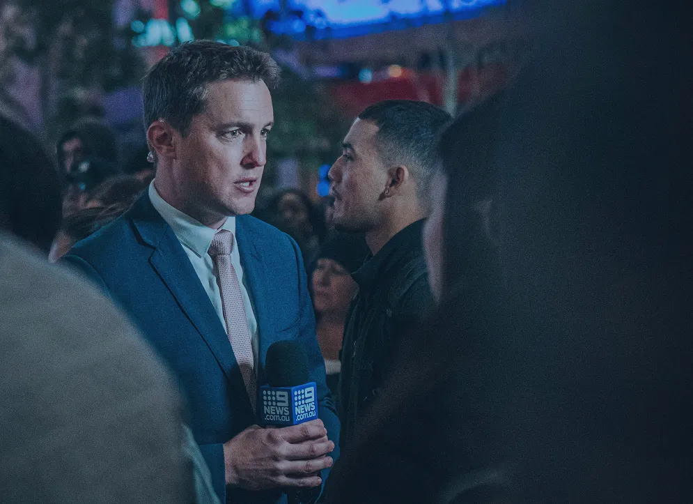 Male news reporter in blue suit holding a 9 News microphone, speaking amid a crowd at night.