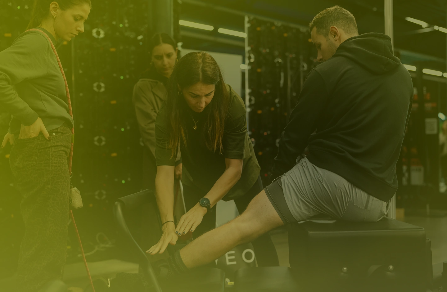 Personal trainer assisting a man stretching his leg while two women observe in a gym setting.
