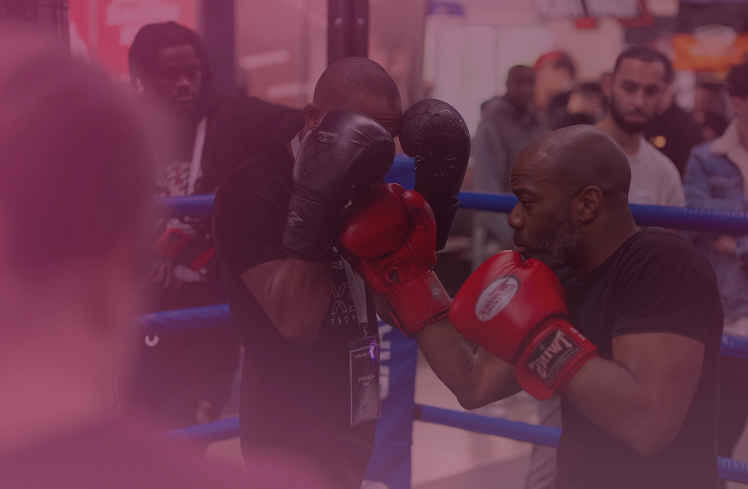 Boxer in red gloves training by practicing punches on a padded mitt held by a coach in a boxing ring with spectators in the background.