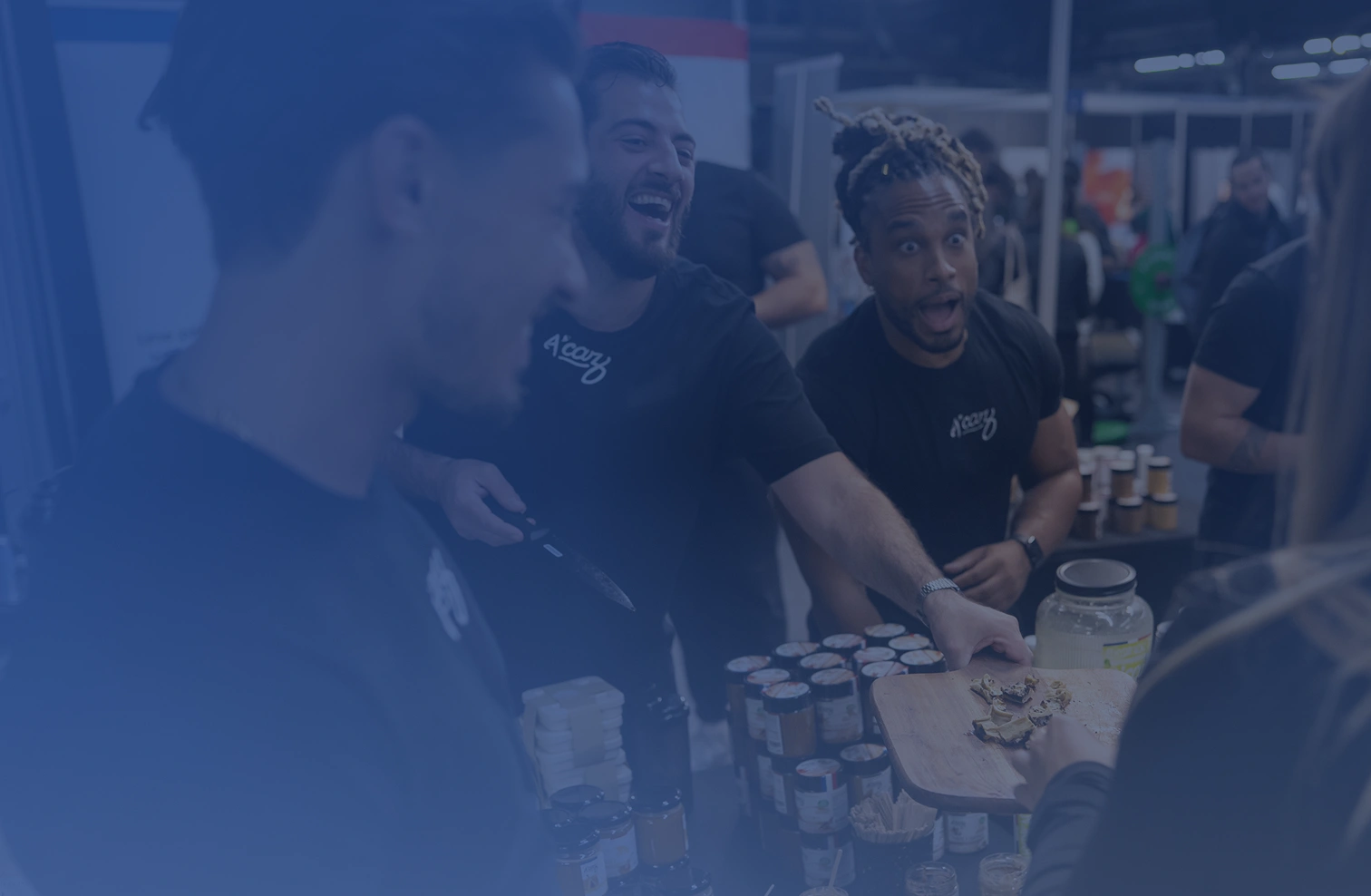 Three men at a market stand offering food samples to a customer, with jars and containers on the table.