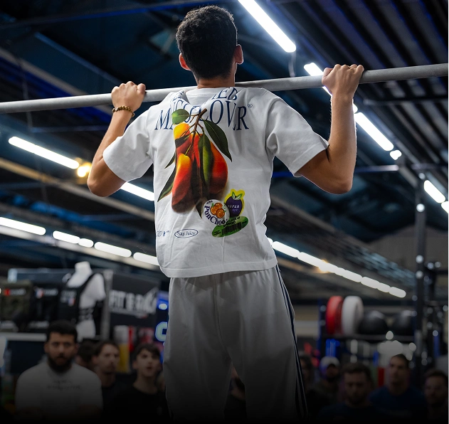 Man in white graphic t-shirt and light pants performing pull-ups in a gym with spectators watching.