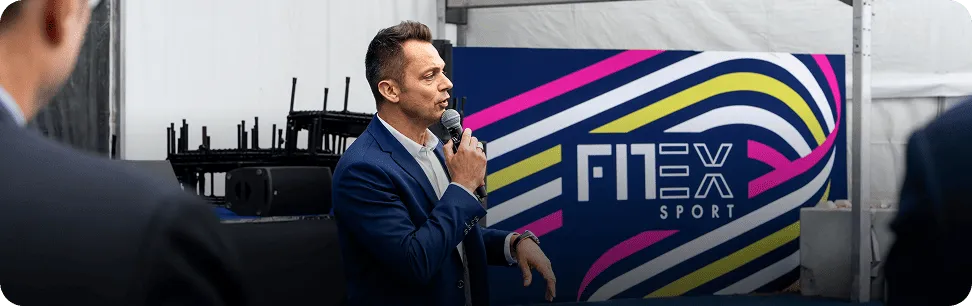 Man speaking into a microphone in front of a colorful FITEX Sport banner at an indoor event.