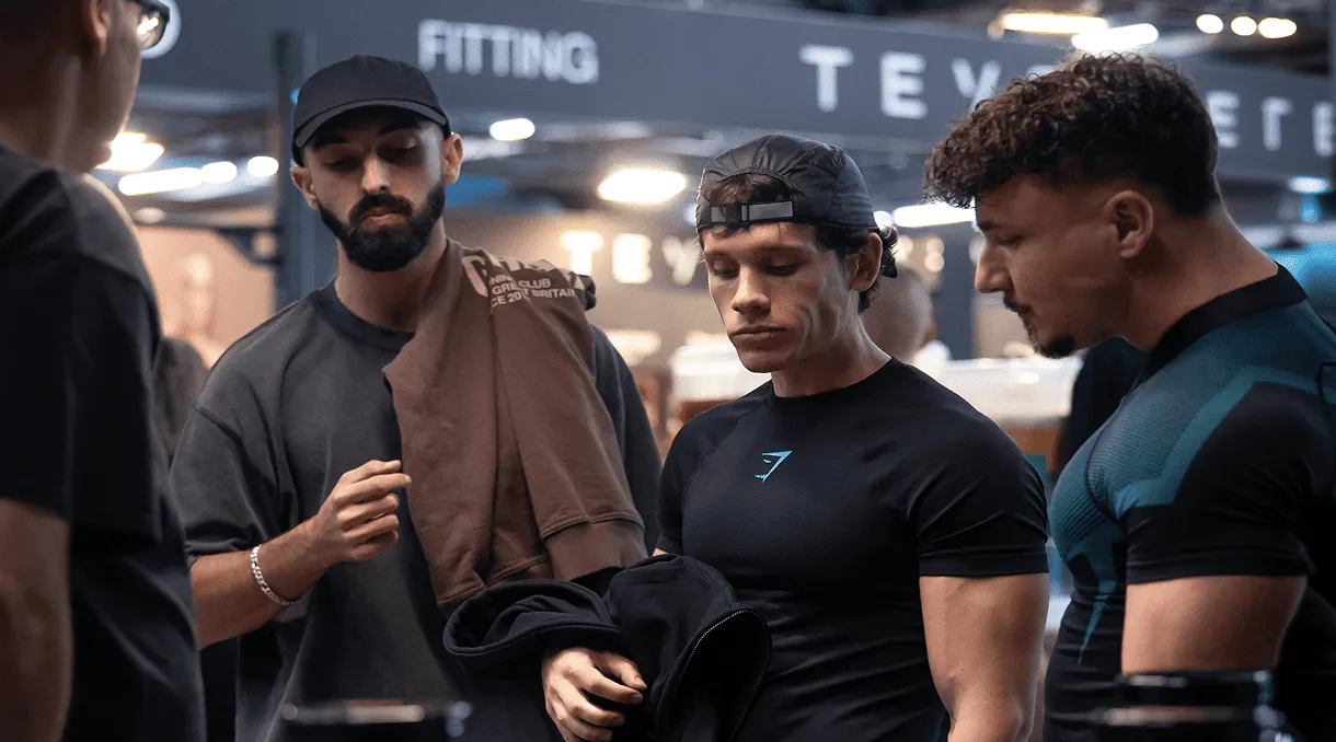 Four young men in athletic clothing standing indoors, with two holding jackets and one wearing a black cap backwards.